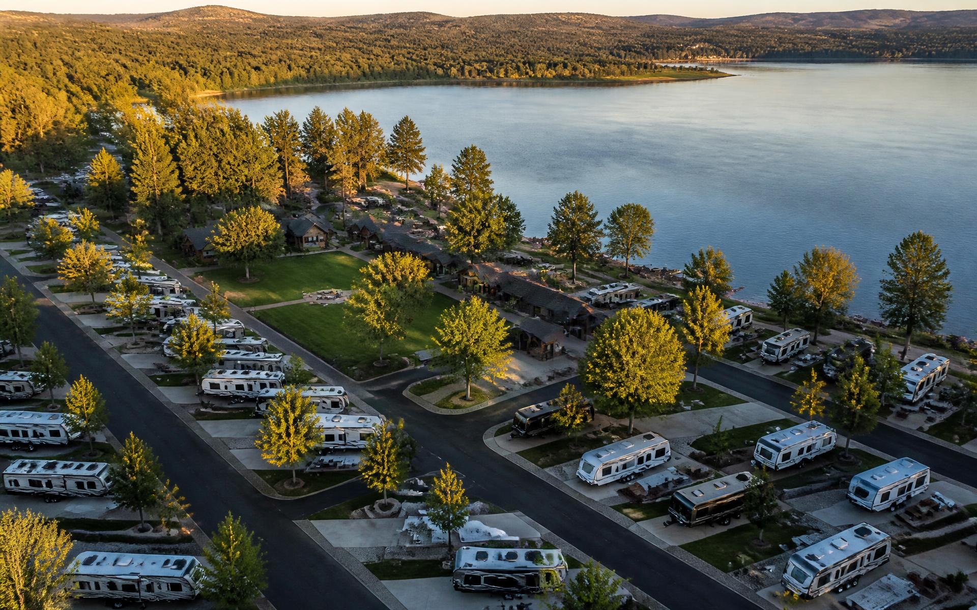 Aerial view of a lakeside RV resort at golden hour with cabins, trees and open lawns